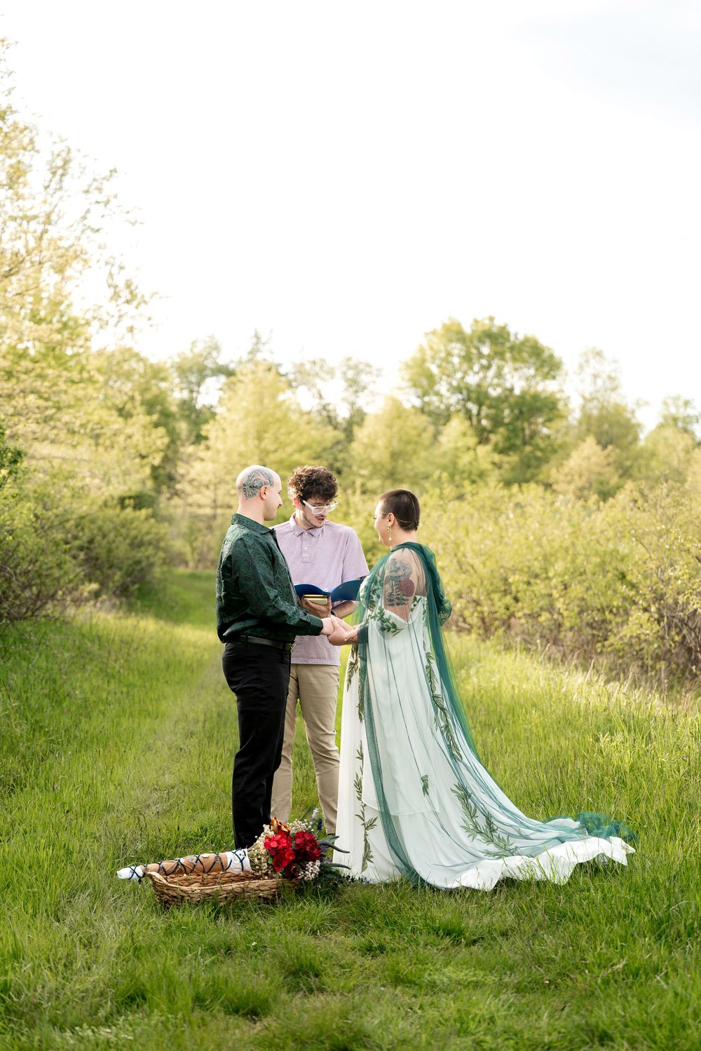 Couple exchanges vows with officiant in a scenic green field surrounded by blooming trees during outdoor elopement.