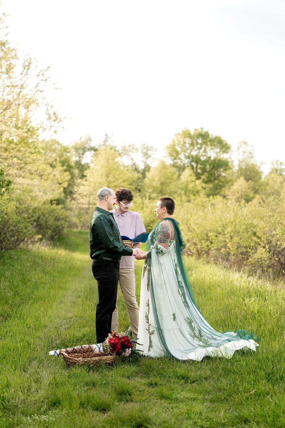 Bride in flowing teal cape gown holds grooms hands during outdoor wedding ceremony in lush green landscape.