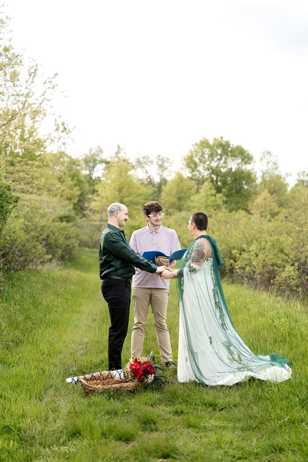 Outdoor wedding ceremony with couple exchanging vows before officiant in lush green meadow, bride wearing teal gown.
