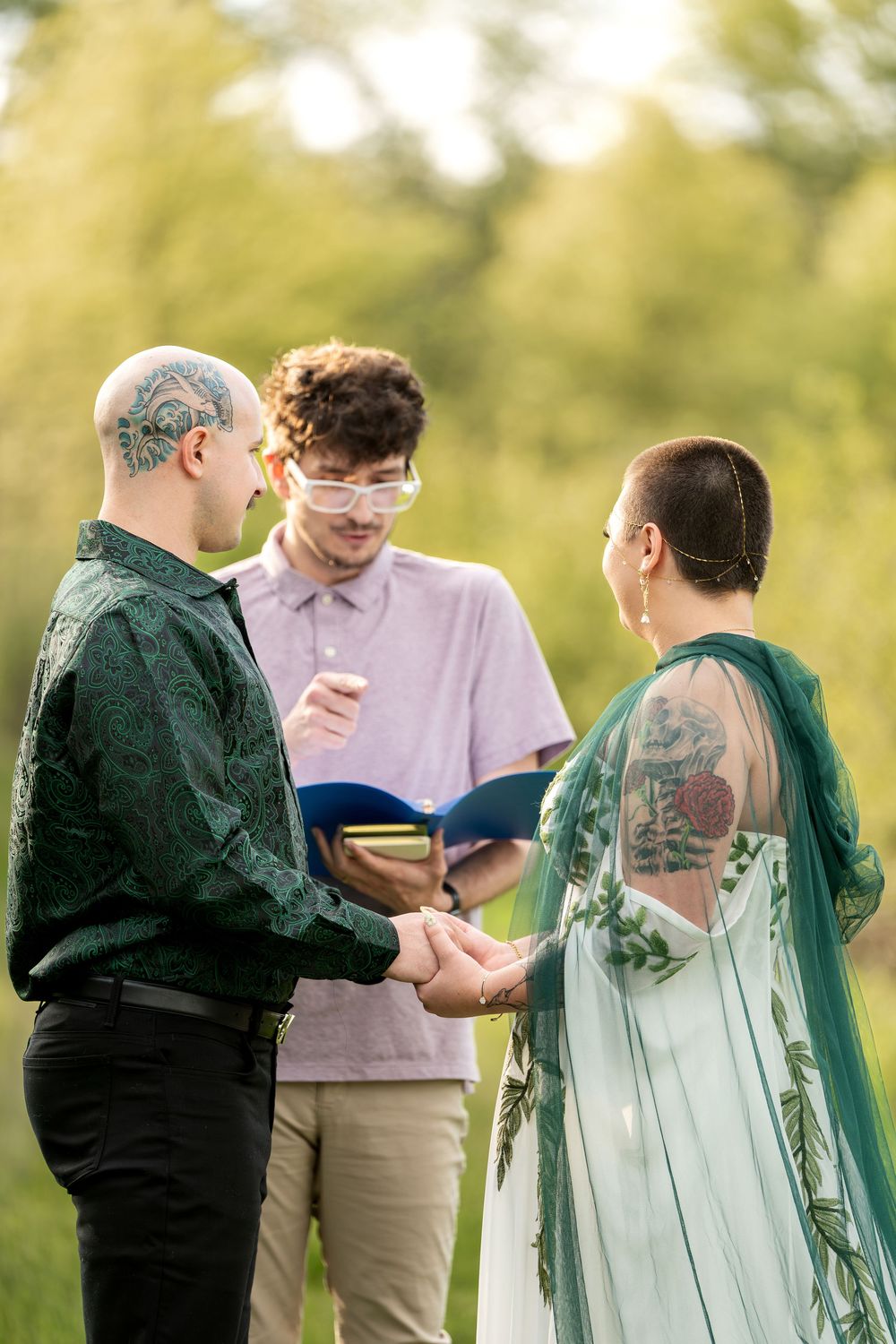 Tattooed groom and bride holding hands during intimate outdoor wedding ceremony with officiant in nature.