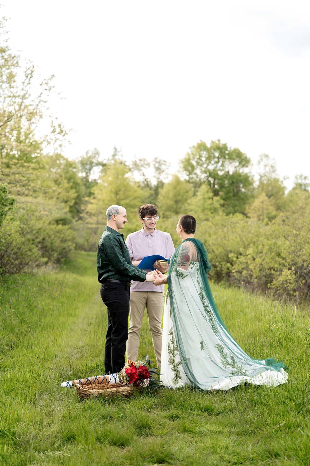 Couple exchanges vows with an officiant in a green meadow, bride in mint cape gown, groom in green shirt.