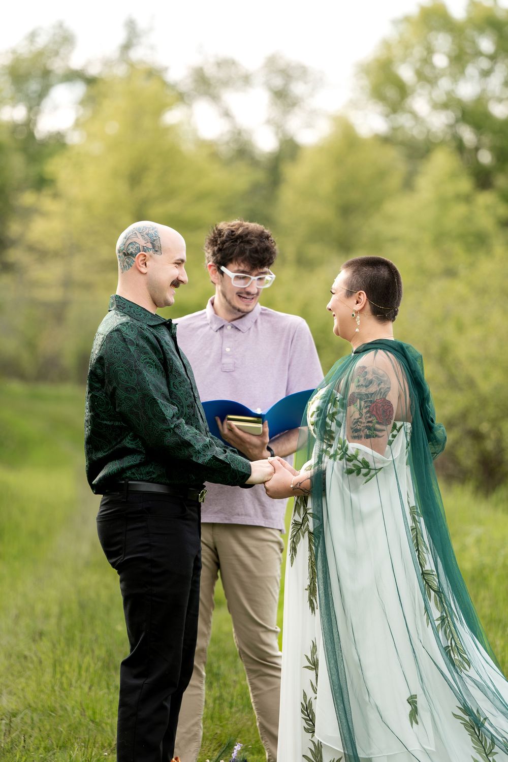 Outdoor wedding ceremony with couple holding hands before officiant holding blue book in green meadow.