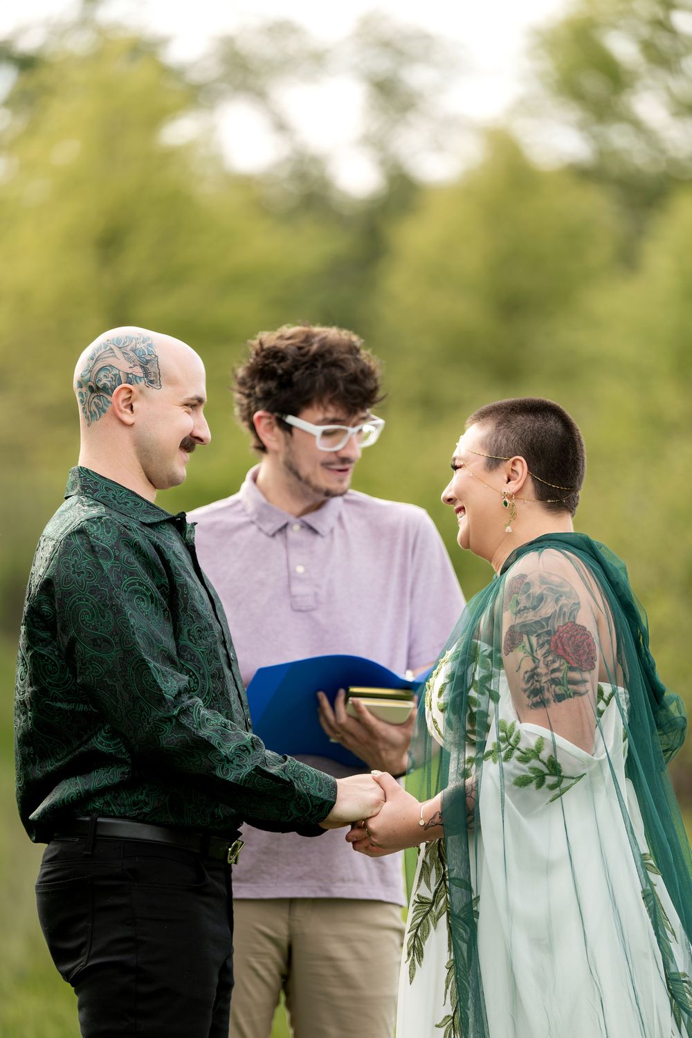 Outdoor wedding ceremony with couple holding hands before officiant in green botanical attire.