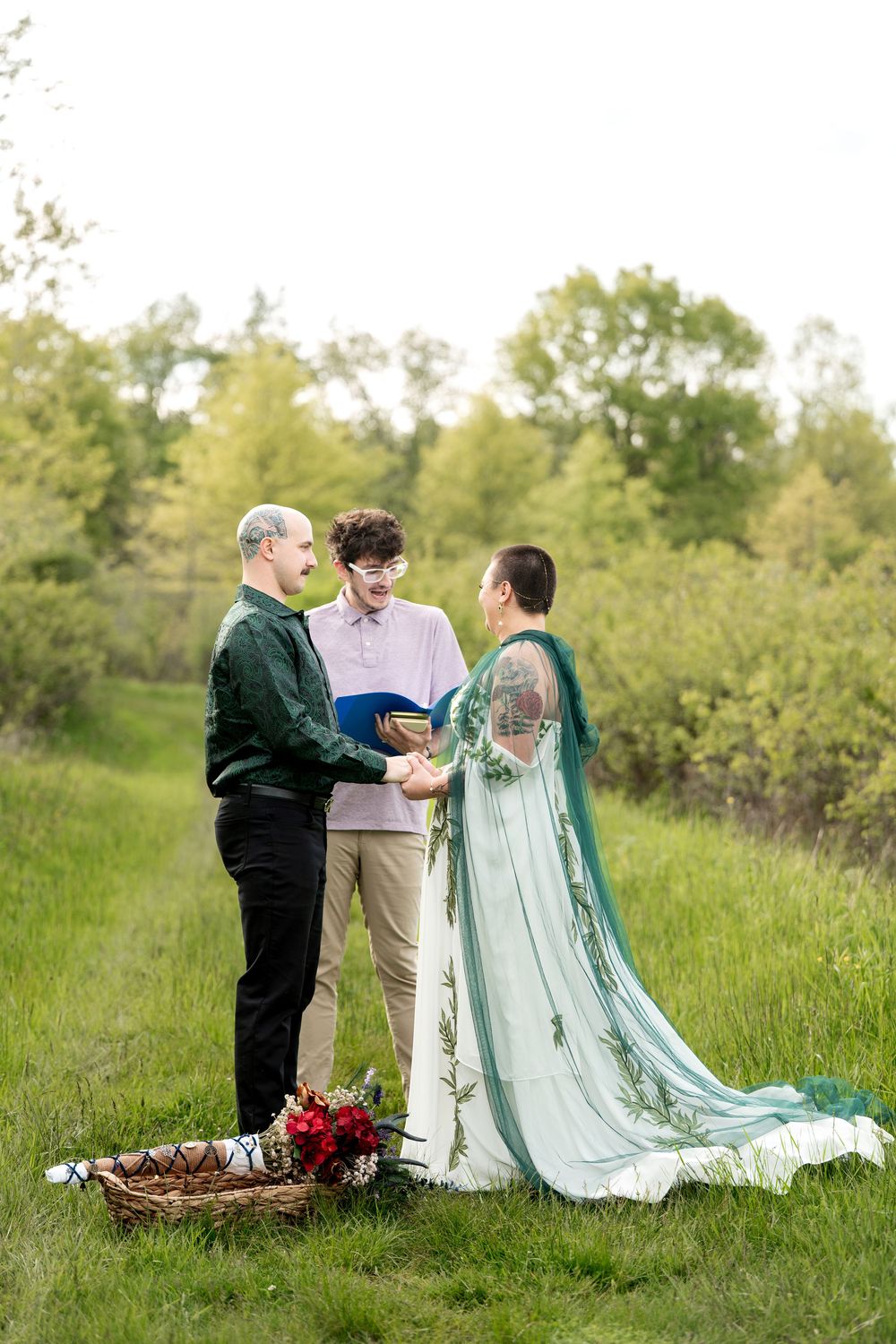 Smiling wedding officiant reads vows as couple holds hands during intimate outdoor ceremony in green meadow.