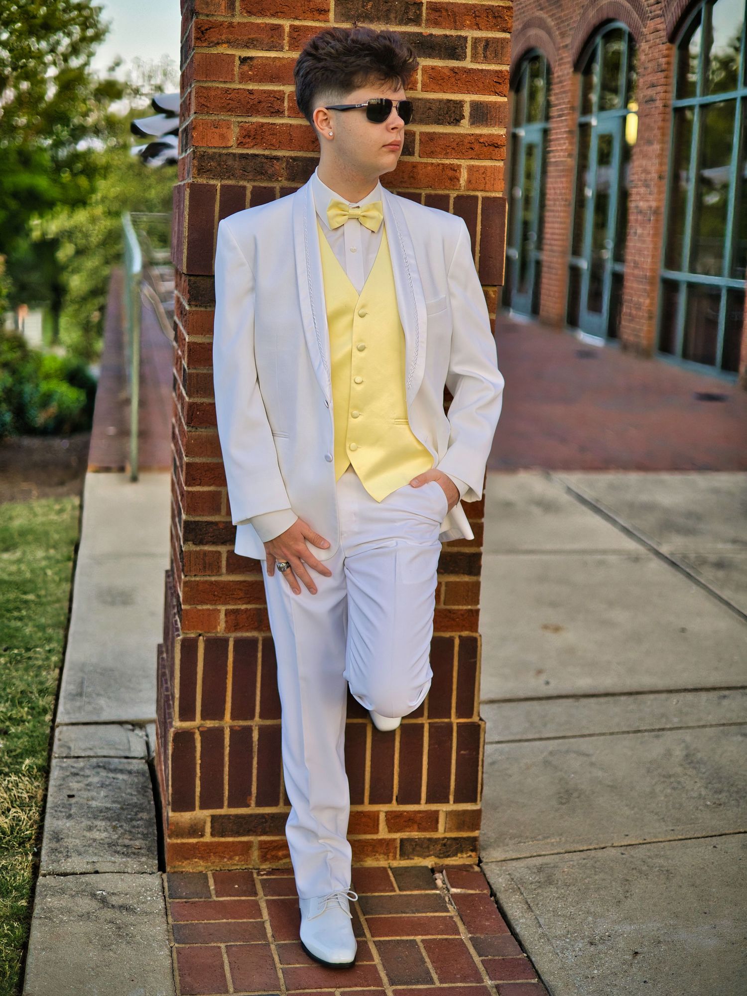 A stylish young man in a white tuxedo with yellow vest and bow tie poses against a brick wall outdoors.