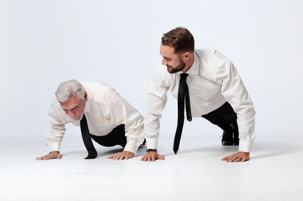 Two men in white dress shirts and black ties doing push-ups on a white floor smiling at each other.