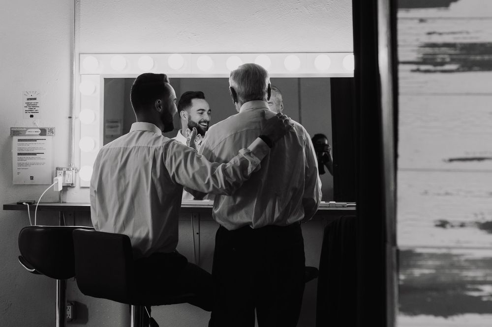 Black and white photo of two men at a mirror in a backstage dressing room setting.