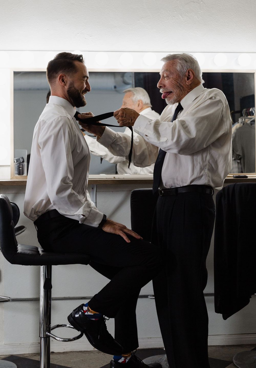 An older man helps a younger man tie his necktie in a dressing room mirror setting.