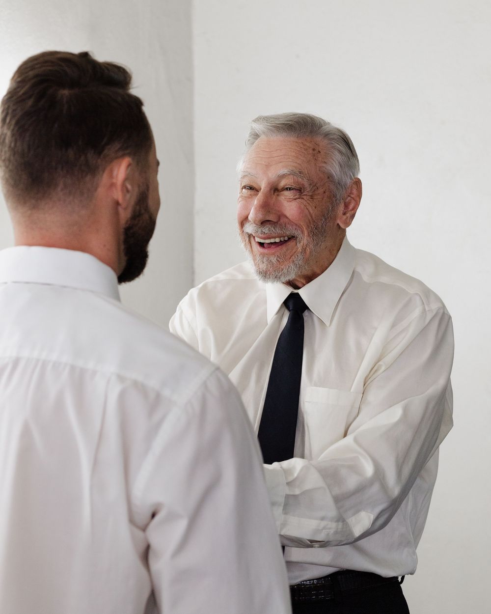 Elderly man in white shirt and black tie smiling warmly while shaking hands with younger bearded man.