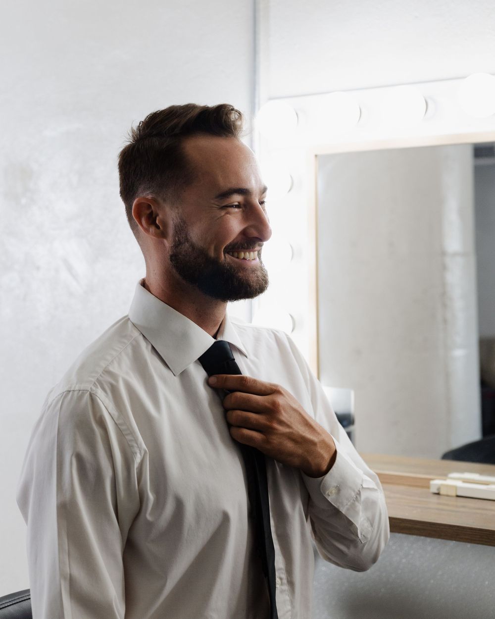 Smiling young businessman adjusting his black tie in front of a mirror in a bright dressing room.