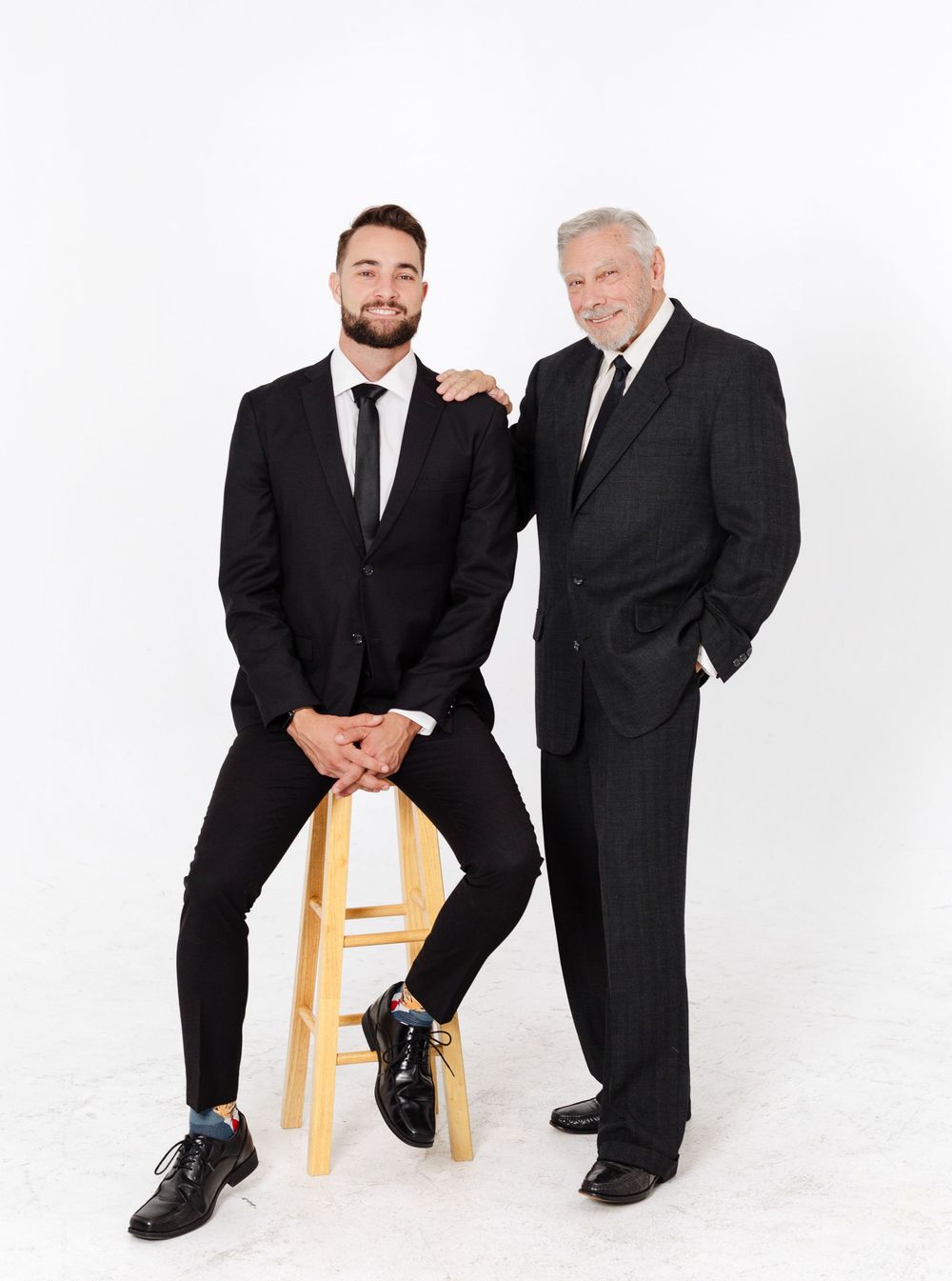 Young bearded man sits on stool smiling beside standing elderly man in black suits against white background.