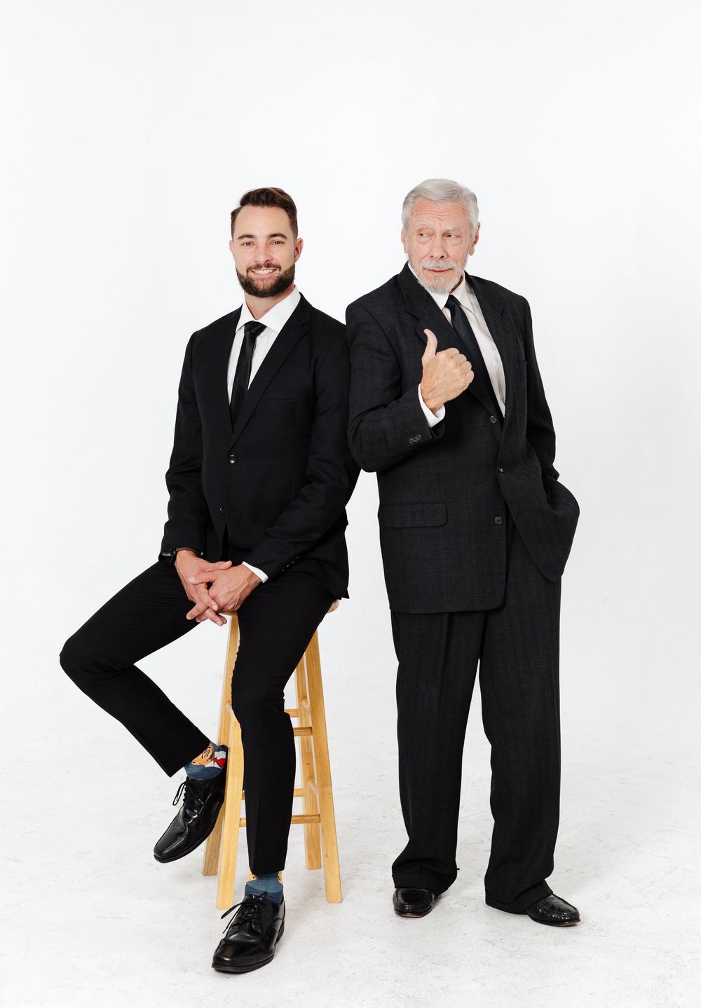 Two men in black suits pose professionally, one seated on a stool and one standing with thumbs up, on white background.