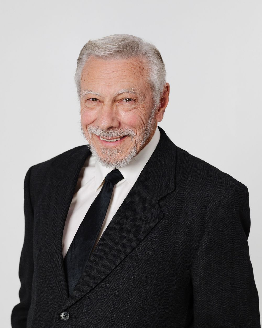 Professional senior businessman with gray beard wearing black suit and tie posing for formal portrait.