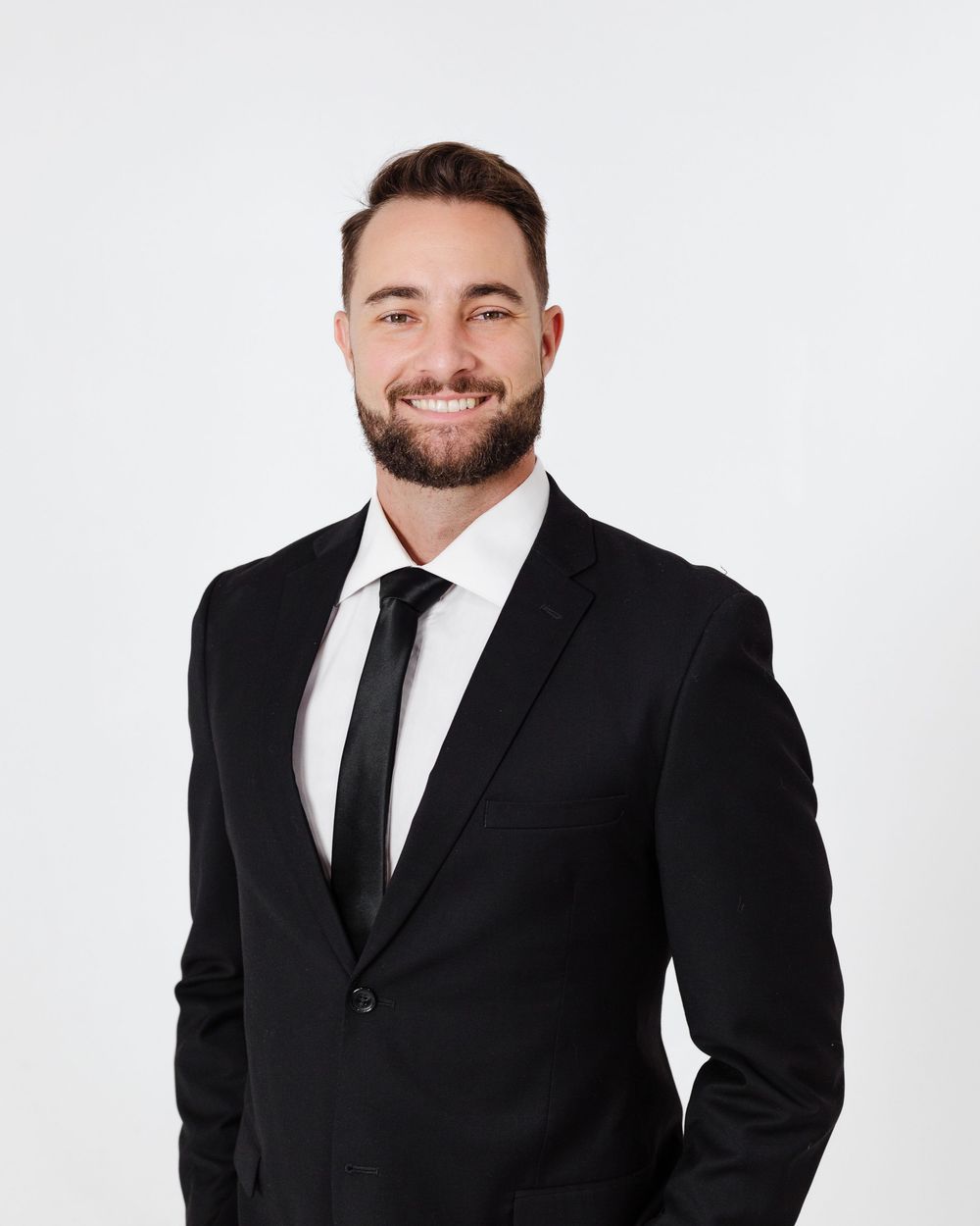 Smiling young professional man in black suit and tie posing for a headshot against white background.
