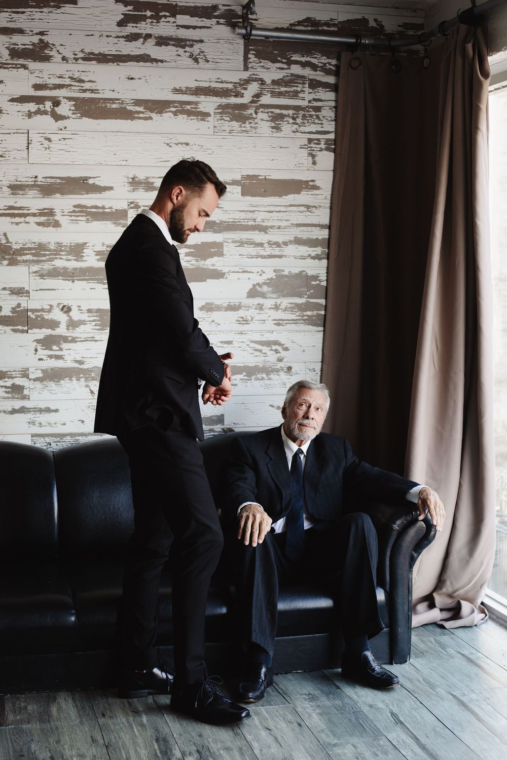 Young businessman in black suit stands over seated elderly man in dark leather chair in rustic room.