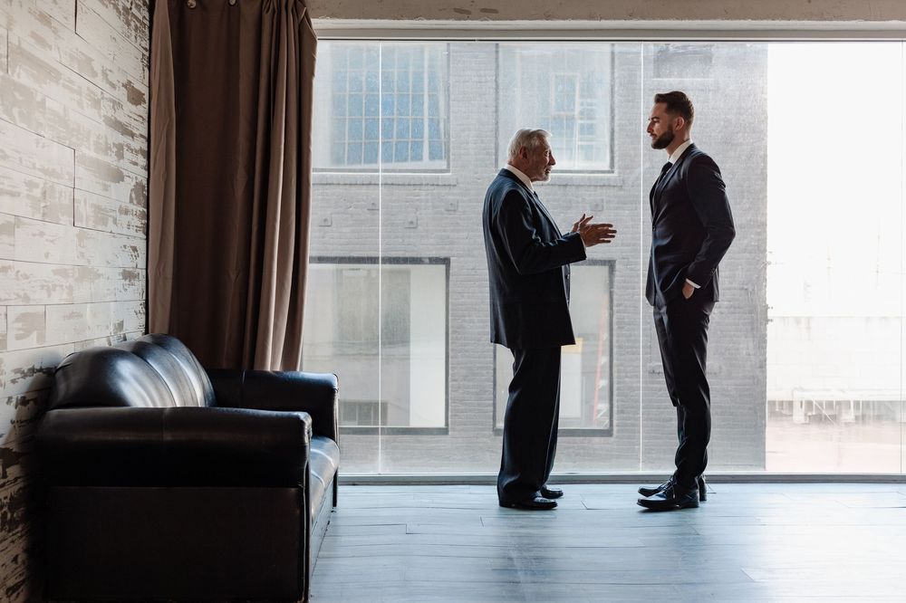 Two businessmen in suits having a professional conversation near large windows in a modern office space.