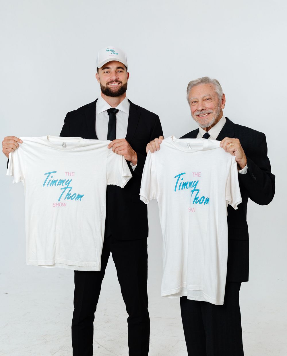 Two men in black suits holding Timmy Thom Show white t-shirts for a promotional photo.