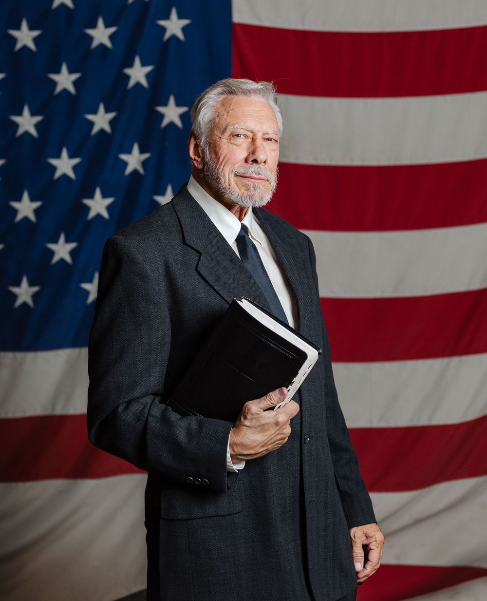 Senior businessman in gray suit holding binder standing before American flag backdrop.