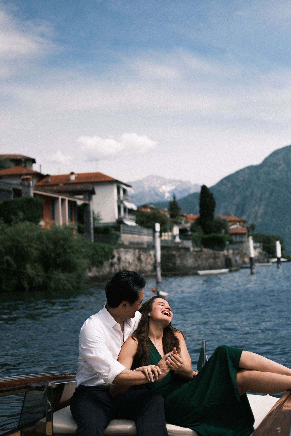 Couple laughing on a boat on Lake Como, Italy, with mountain views and waterfront villas in the background.