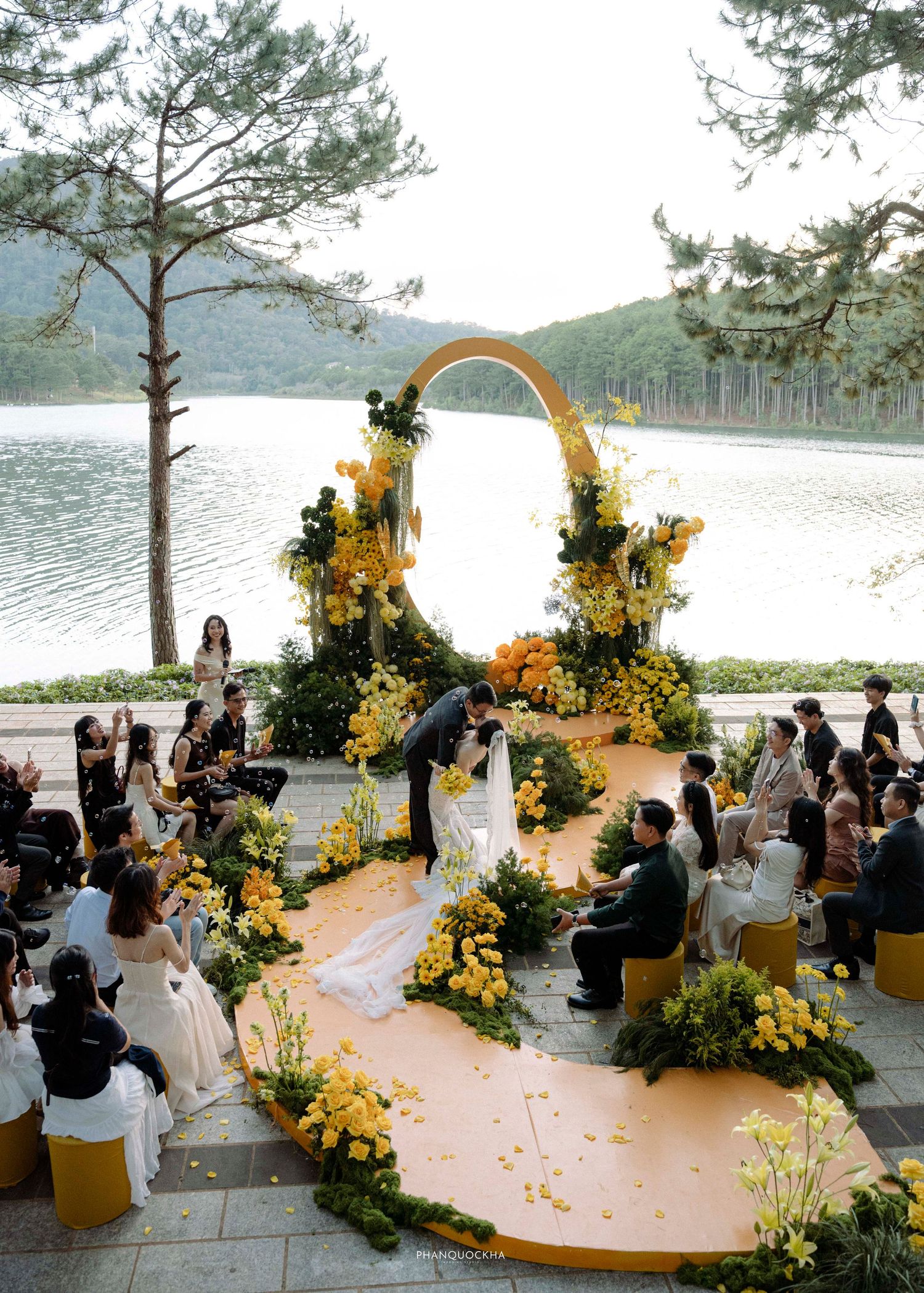 Elegant lakeside wedding ceremony with yellow floral arch and orange aisle runner.