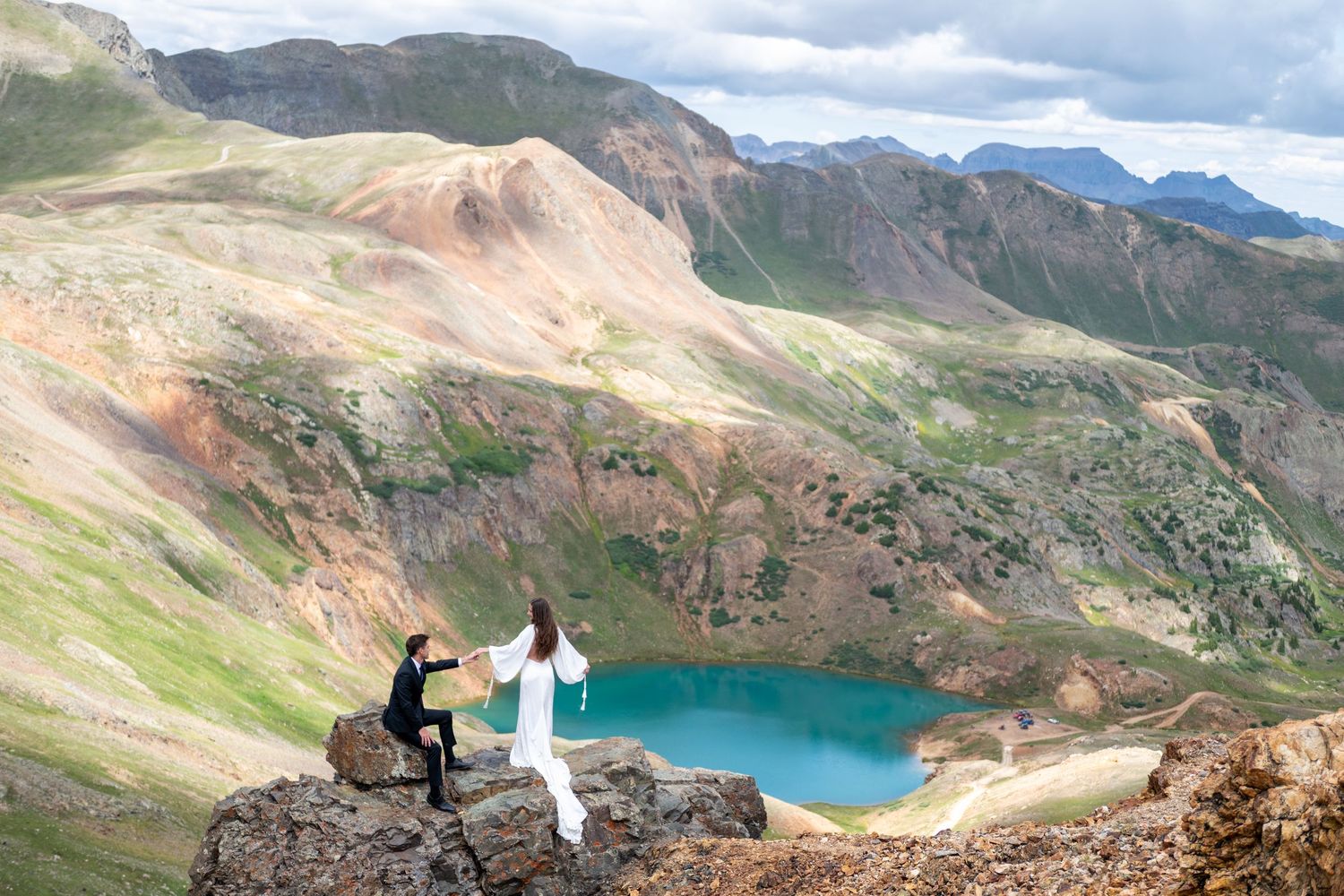 An eloping couple sitting in wooden chairs admiring snow-capped mountain views in a scenic alpine meadow with evergreen trees.