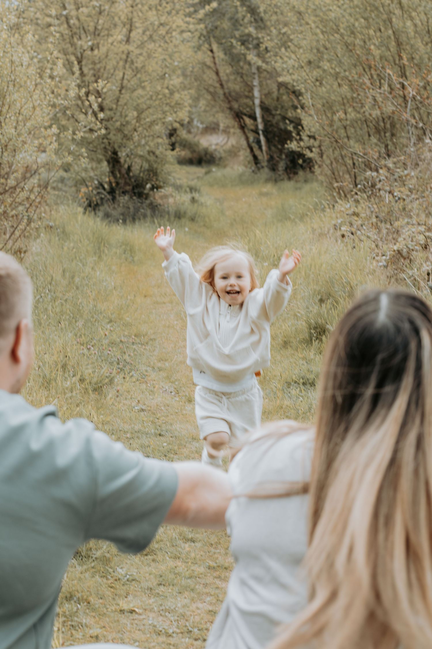 A joyful toddler in white runs with arms raised toward parents in a sunny meadow surrounded by green trees.