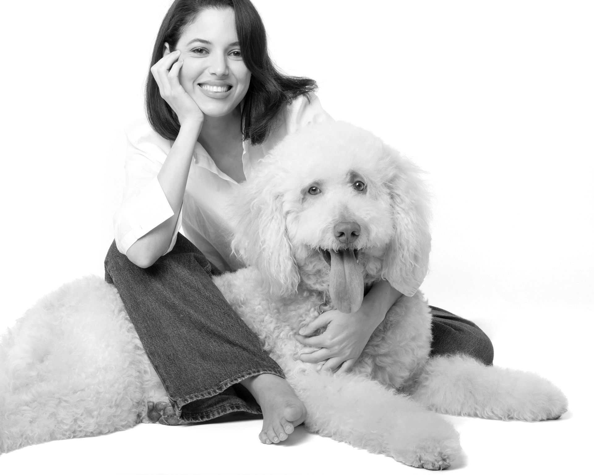 Black and white photo of a smiling woman posing with a fluffy white Goldendoodle dog on a white background.