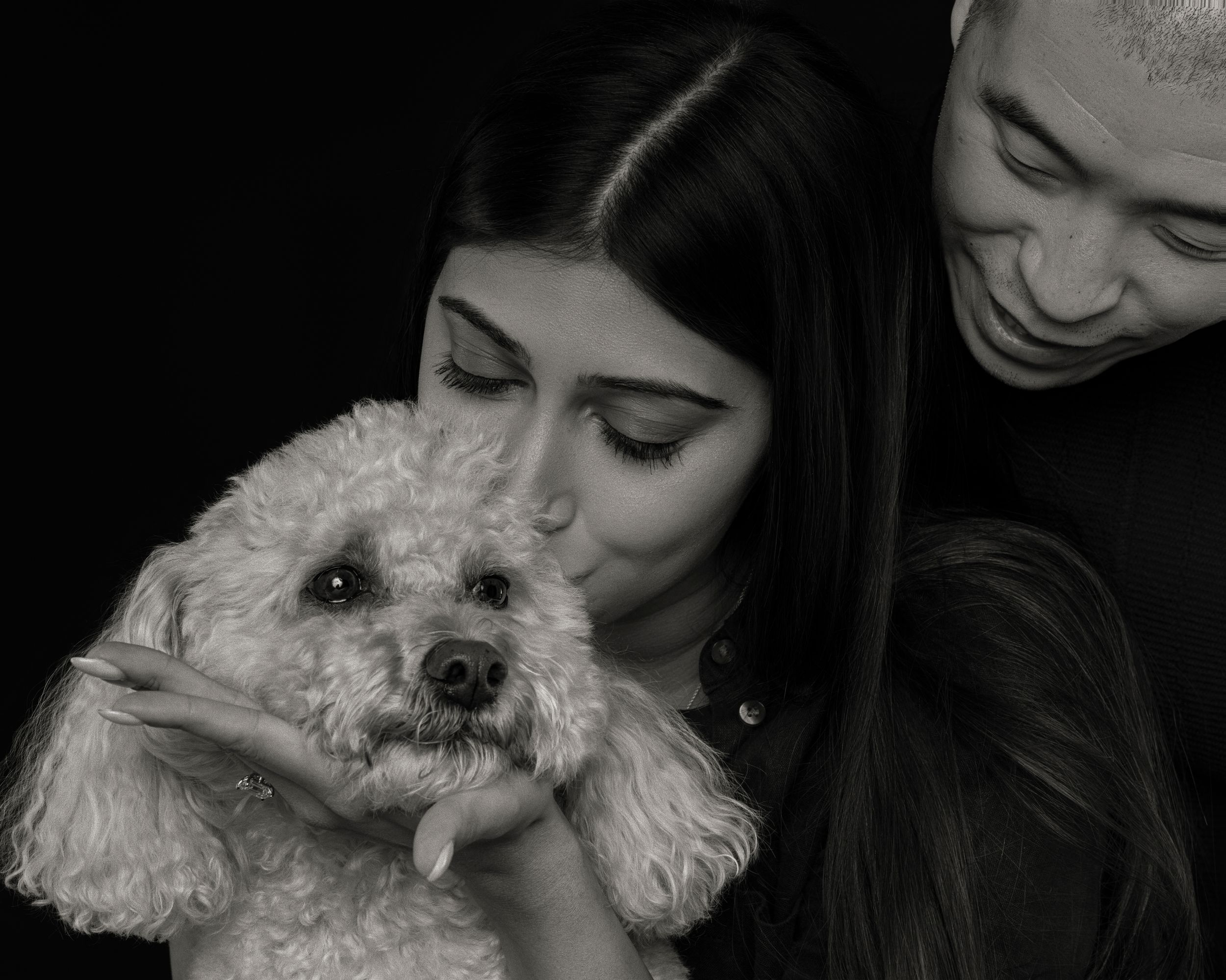 Black and white photo of a couple lovingly holding and kissing a fluffy white poodle dog.
