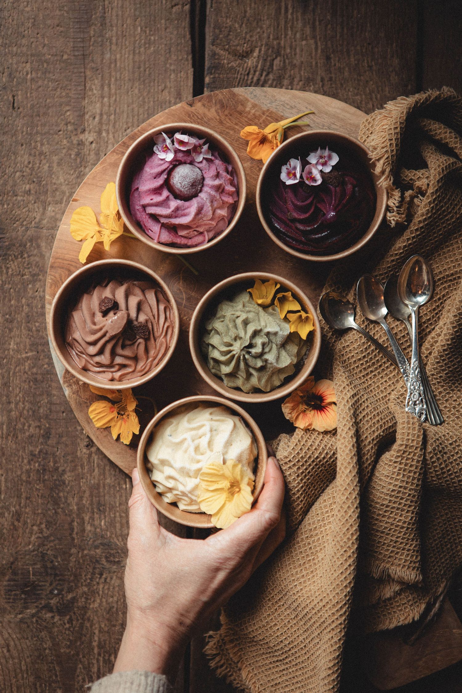 Hand holding a bowl of white ice cream surrounded by colorful frozen yogurt bowls on a wooden board with flowers.