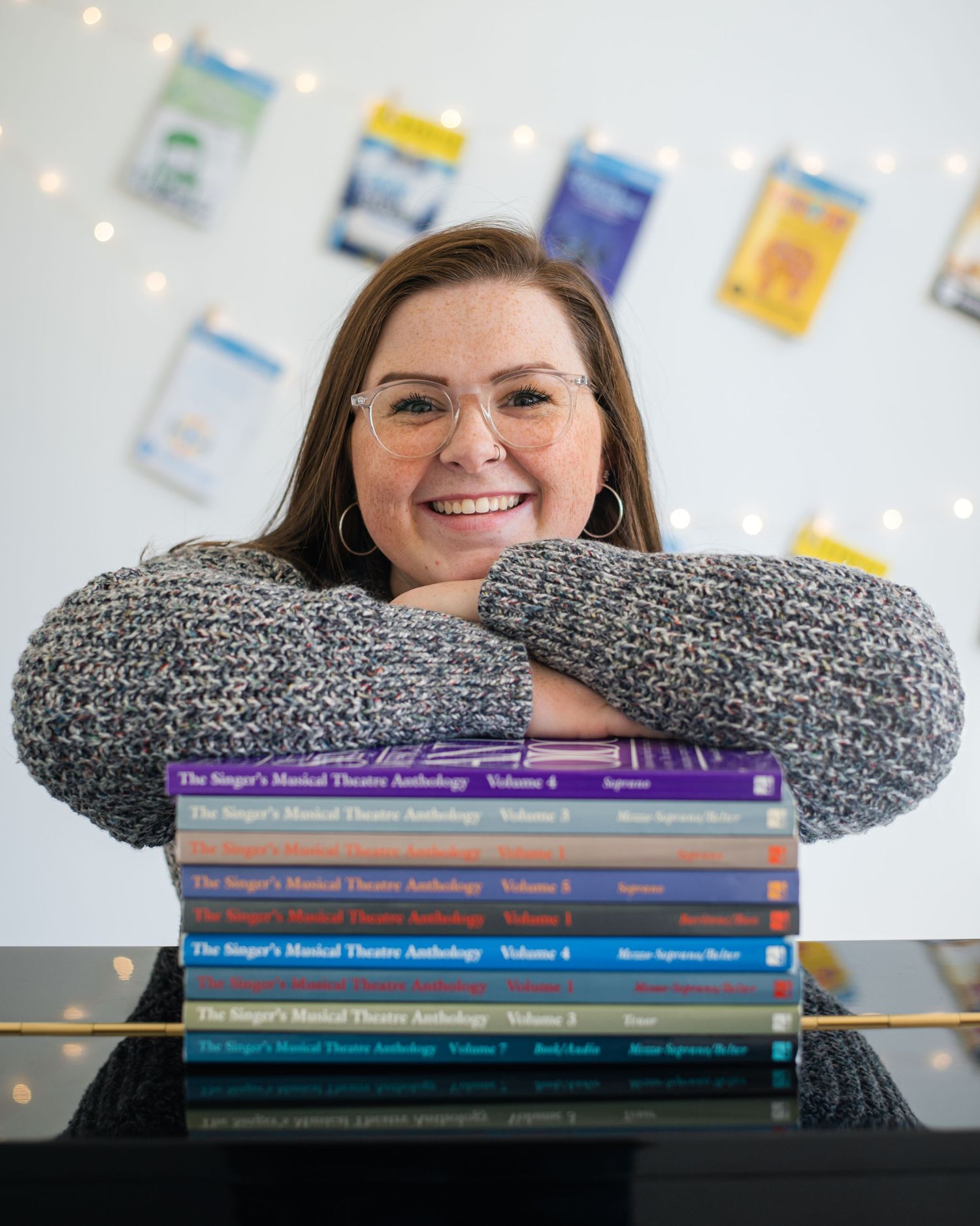 Woman smiling behind a stack of Singer's Musical Theatre Anthology books on a piano in a bright studio.