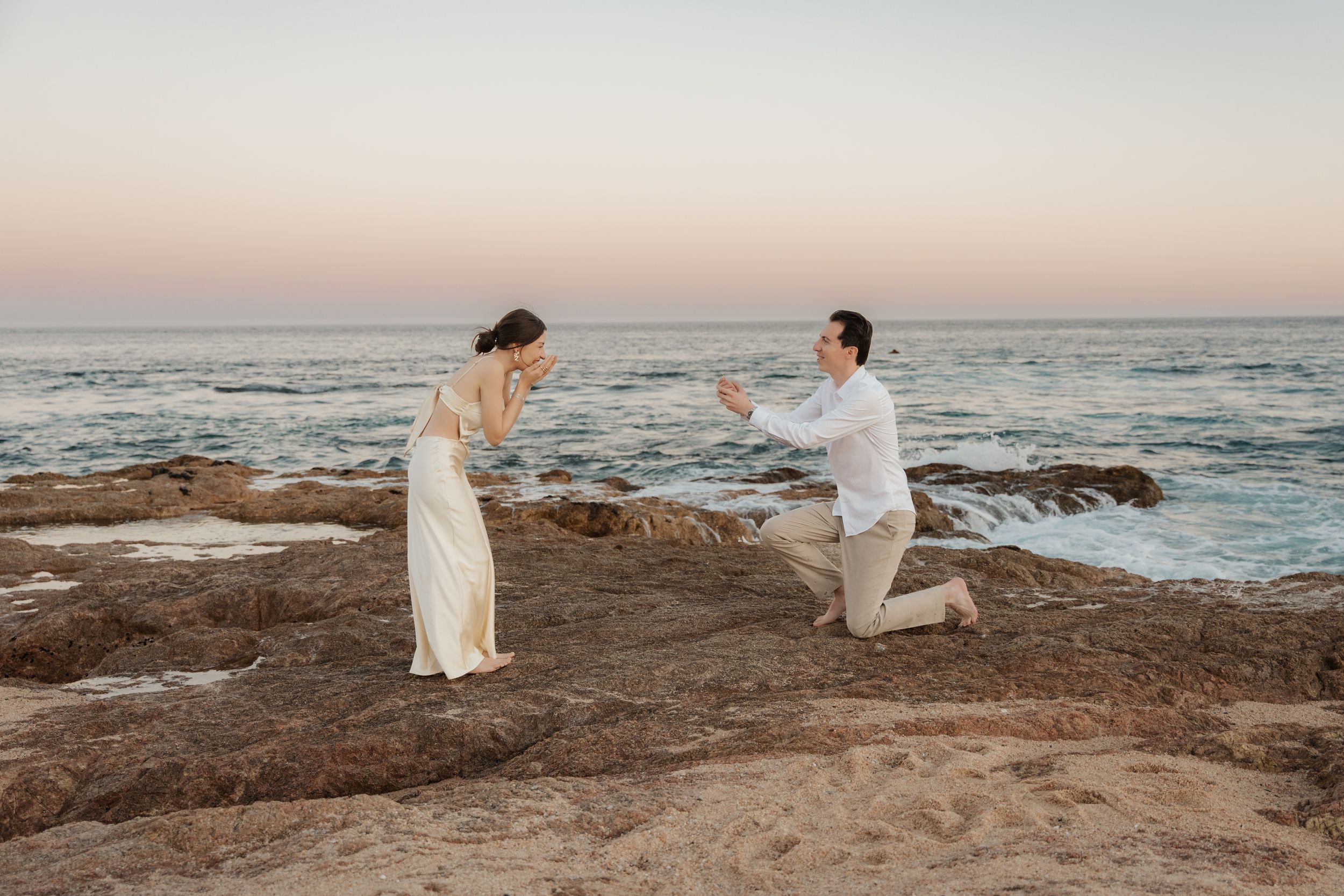 Man proposes to woman on rocky ocean cliff at sunset, both dressed elegantly in white.