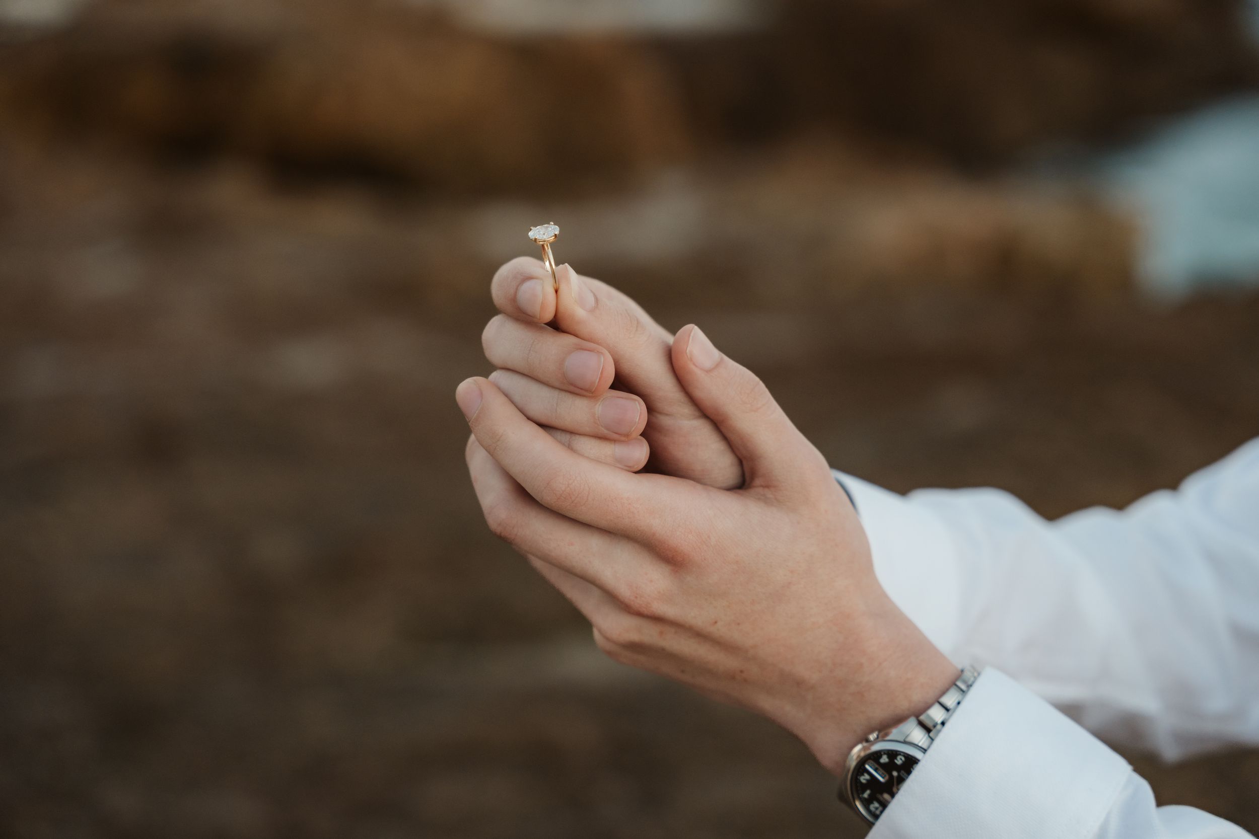 Close-up of hands holding a small delicate flower or ring outdoors, wearing a white shirt and silver watch.