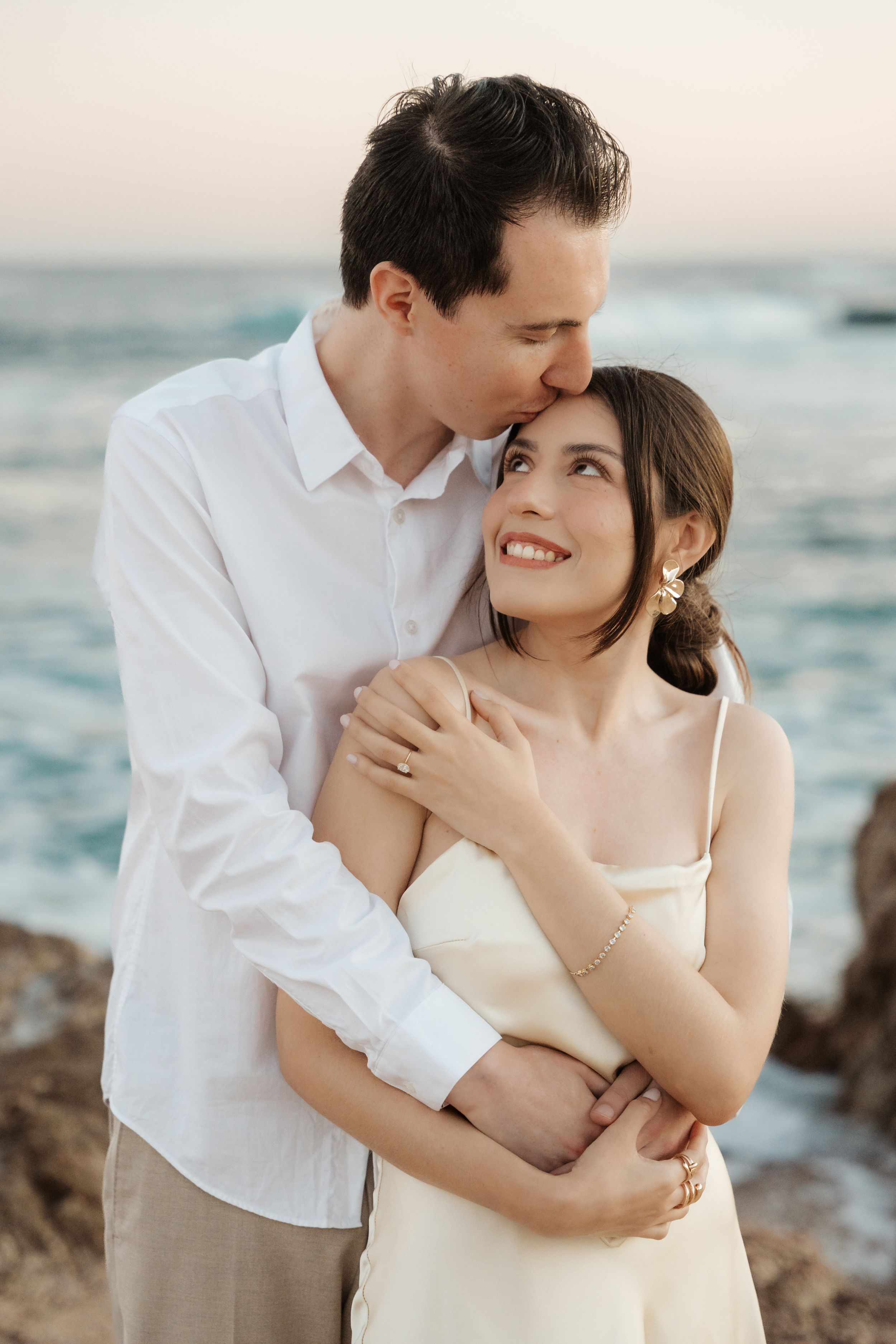Romantic couple embracing at sunset by the ocean, man kissing woman's temple while she smiles, wearing white outfits.