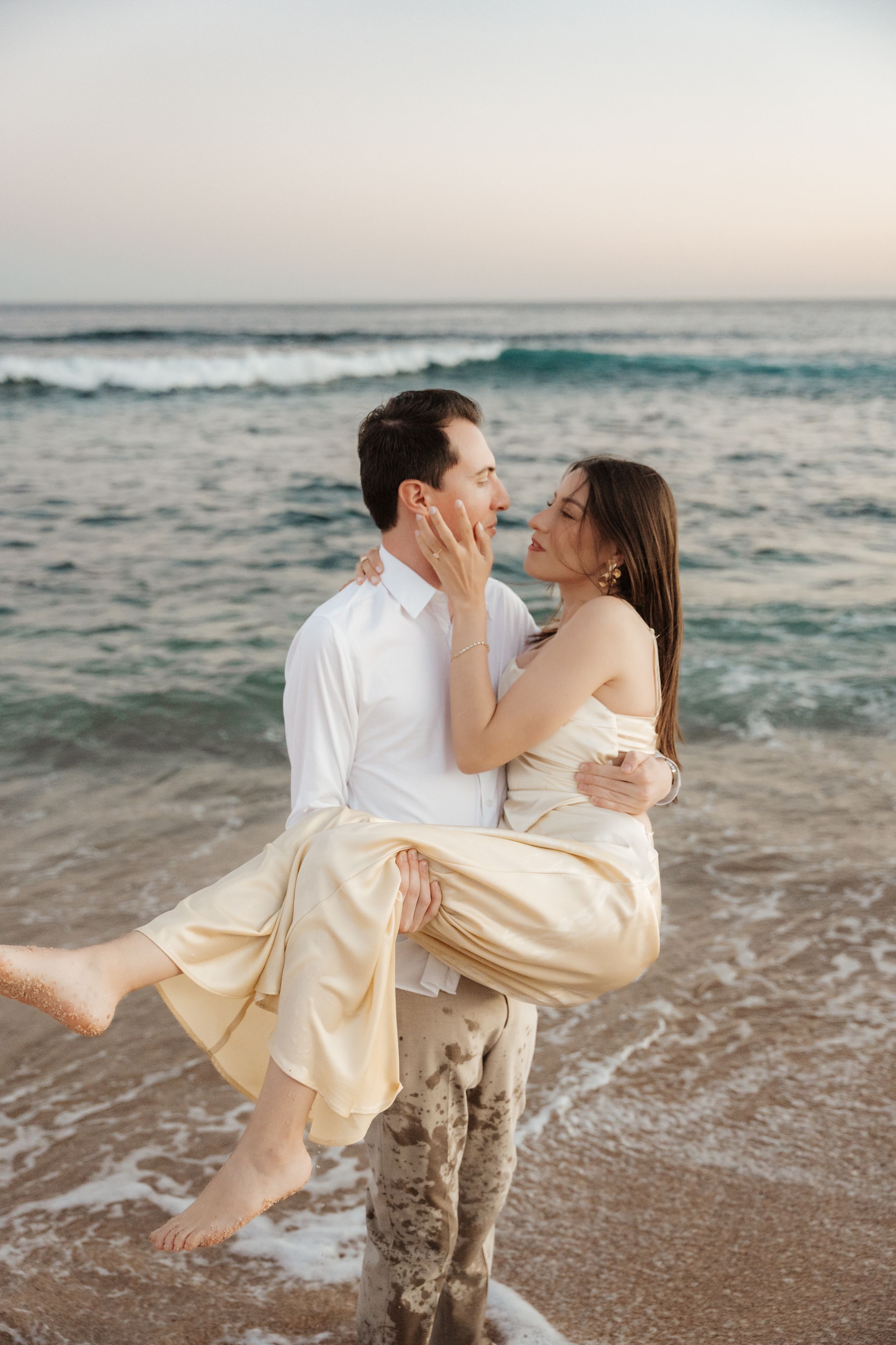 Romantic couple embracing on a beach at sunset, man carrying woman in yellow dress near ocean waves.