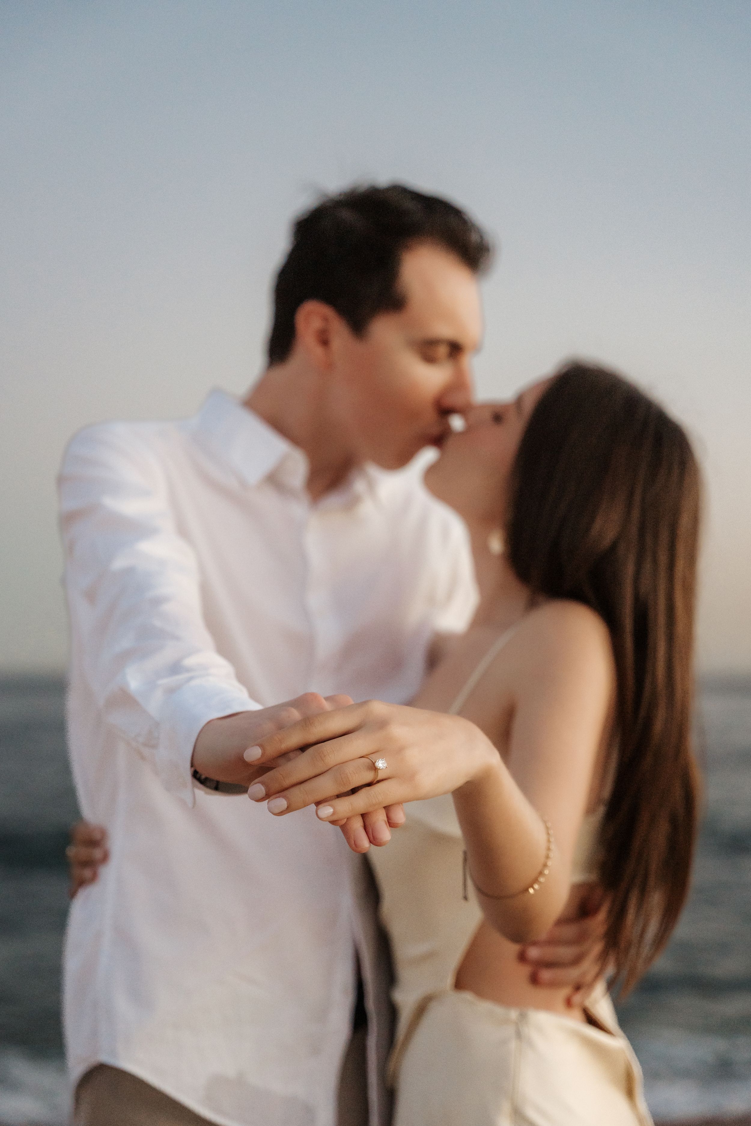 Couple kissing at beach sunset, woman displaying engagement ring toward camera, ocean backdrop.