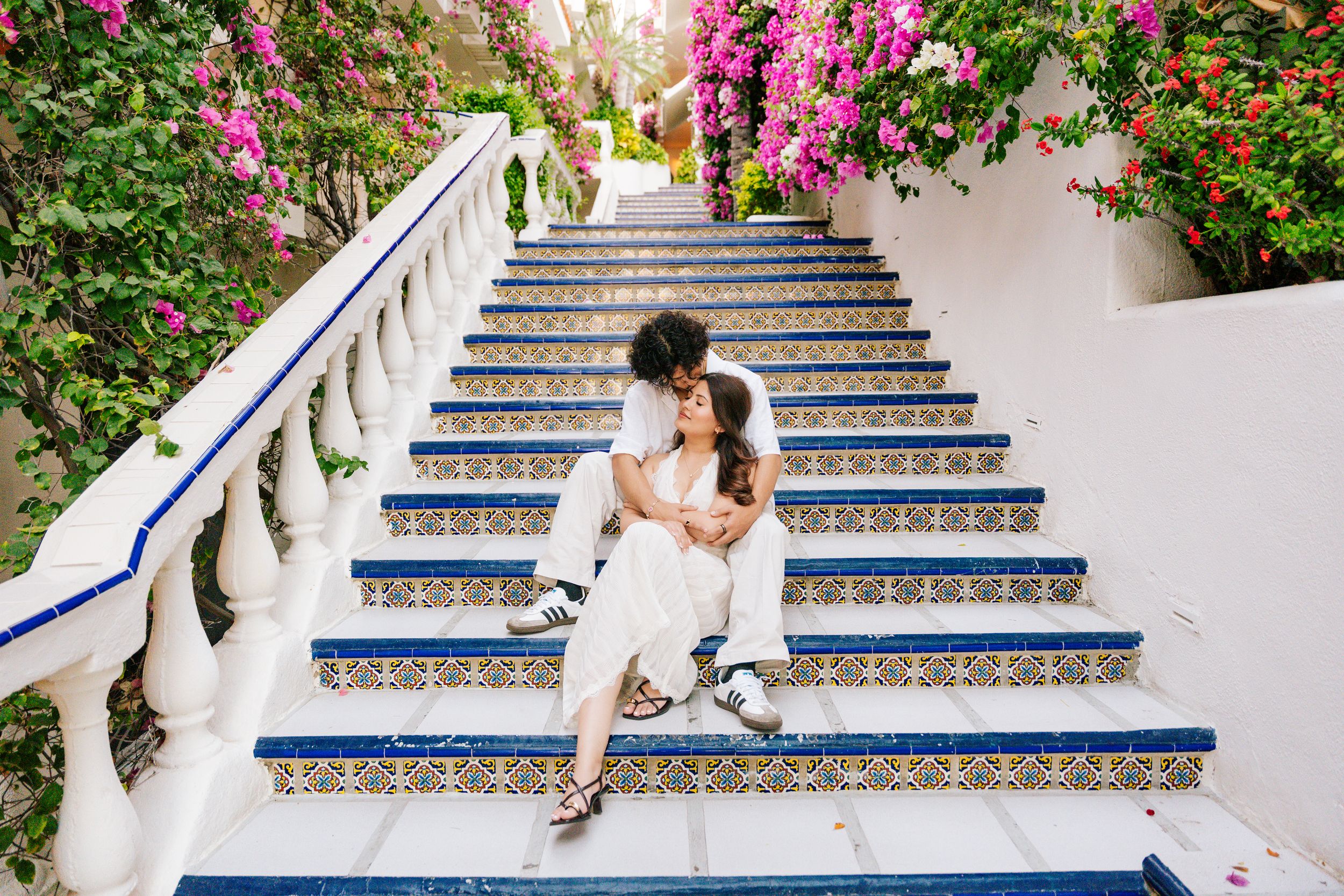 Couple sitting on ornate tiled steps surrounded by pink bougainvillea flowers in a romantic Mediterranean setting.