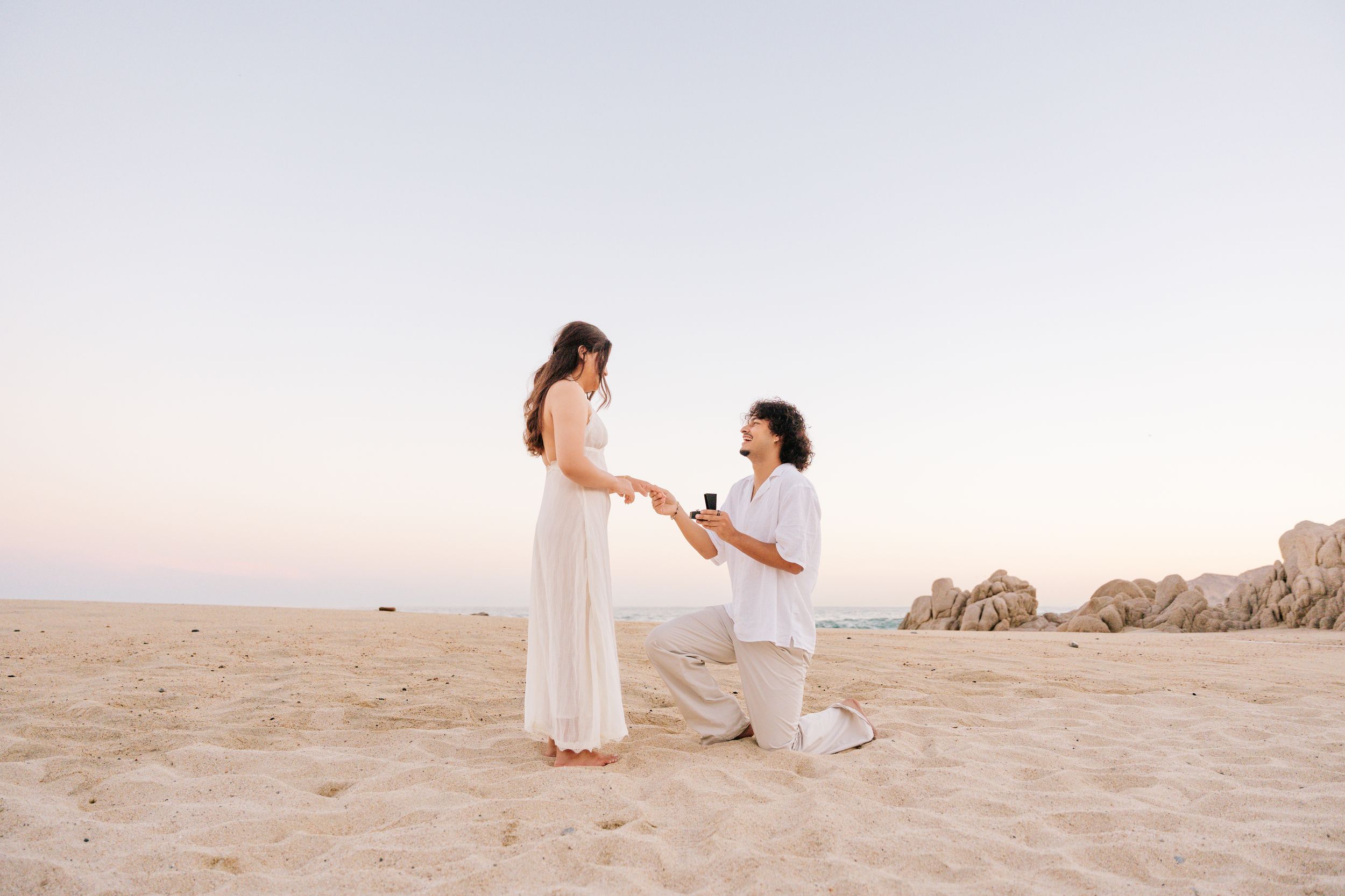 Man kneeling on sandy beach proposing to woman in white dress at sunset with rocky cliffs in background.