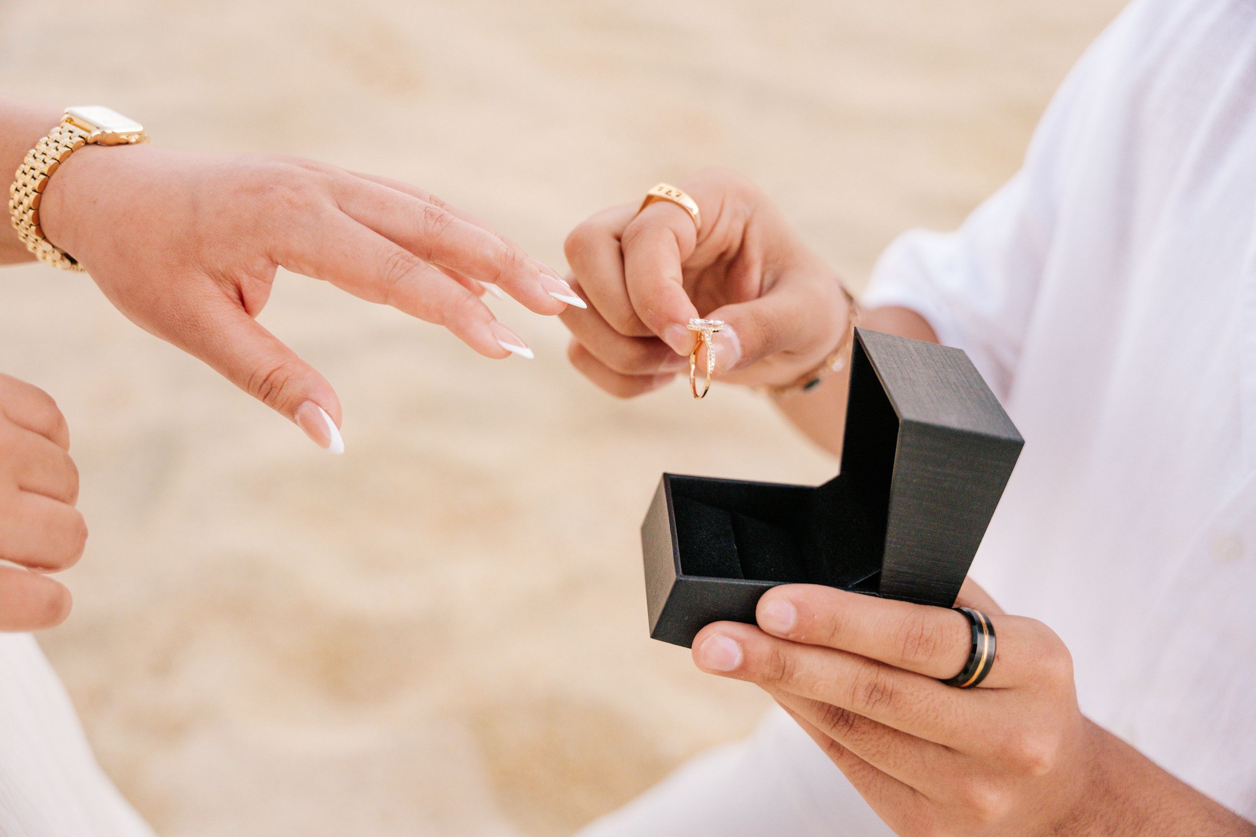 Person placing a gold engagement ring on a partner's finger from a black ring box on a sandy beach.