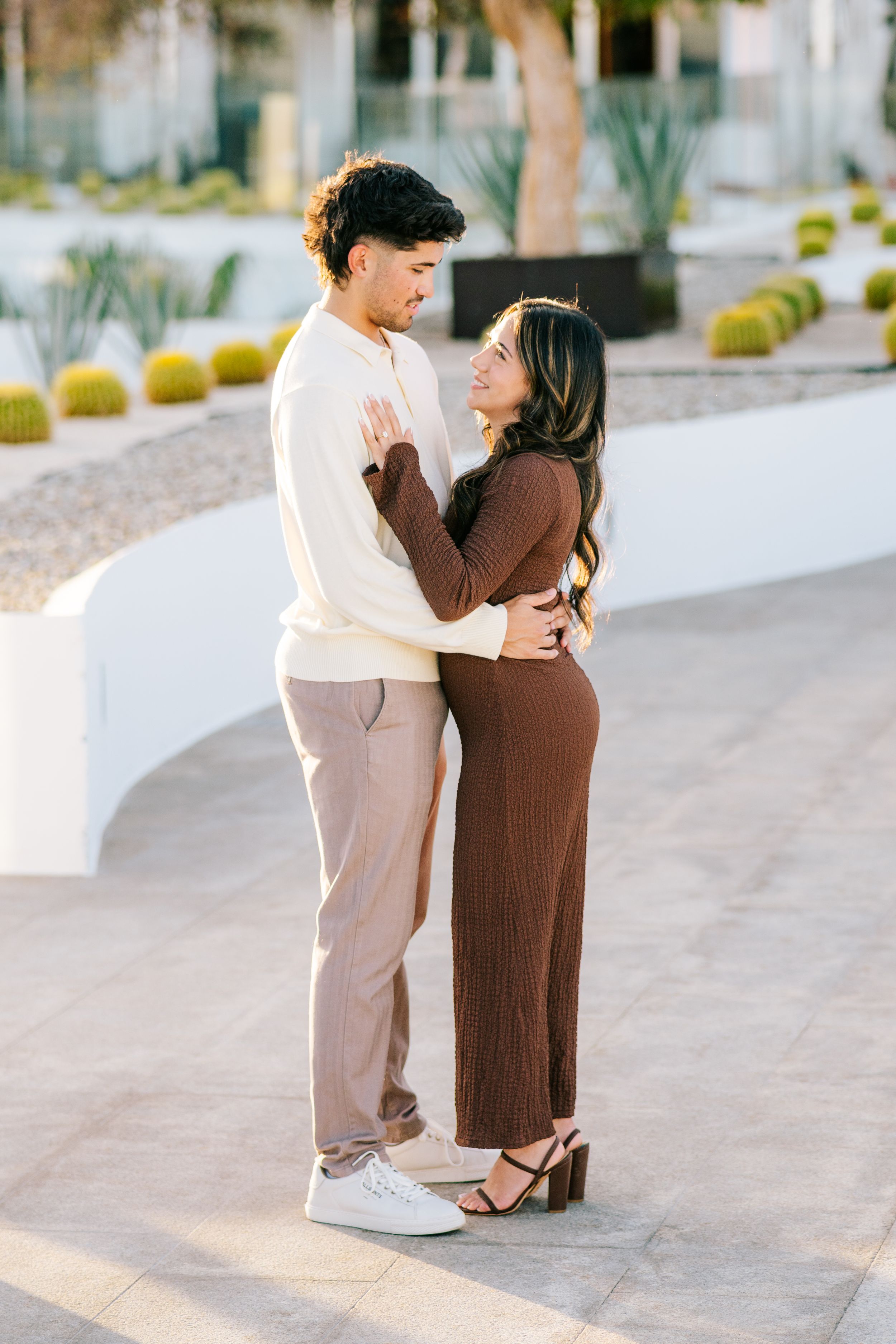 A couple embraces and gazes at each other in a desert garden, wearing neutral earth tones, with cacti in the background.