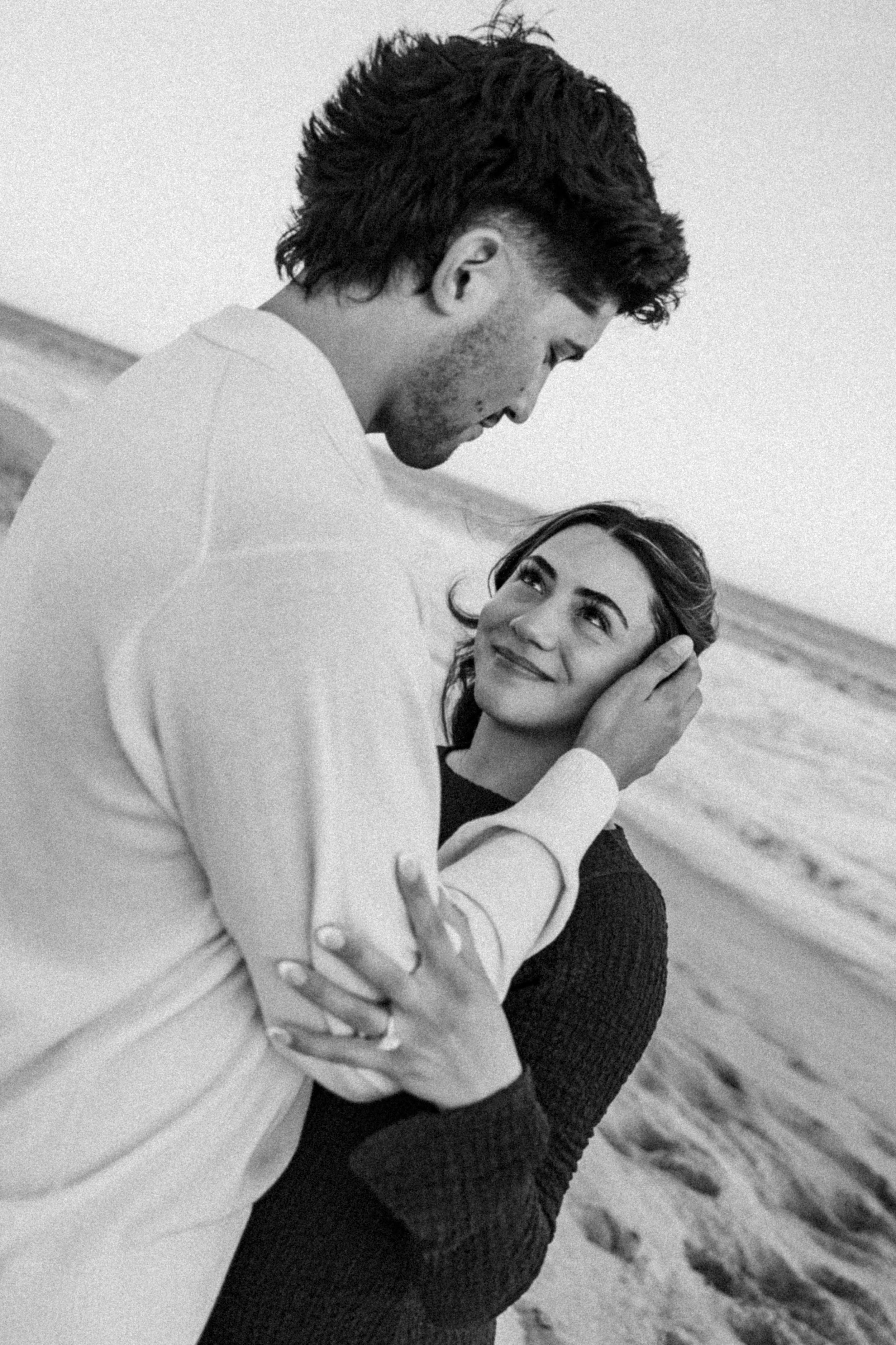 Black and white photo of a couple embracing on a beach, woman smiling up at man tenderly holding her face.