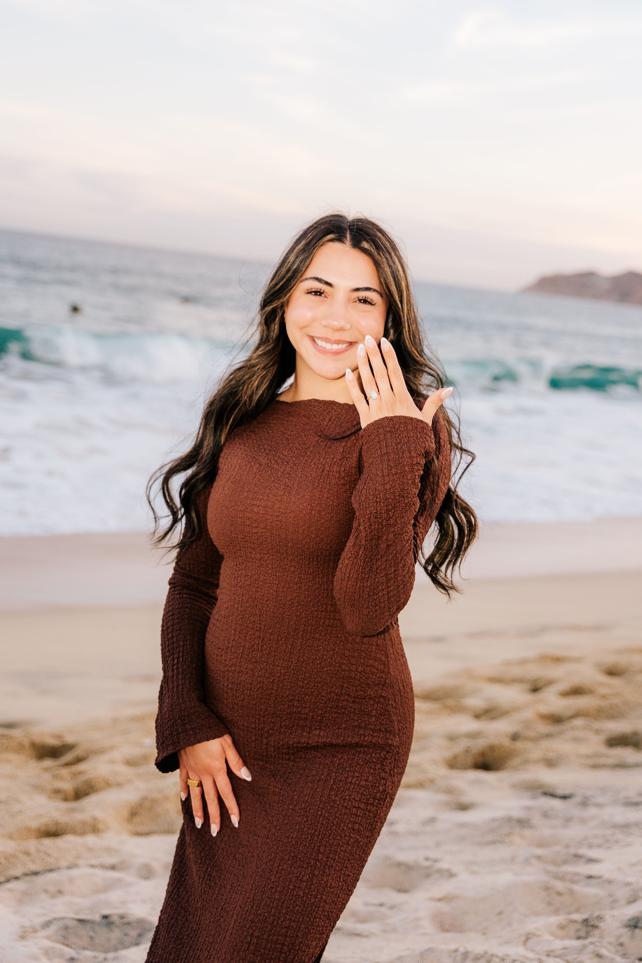 Smiling pregnant woman in brown ribbed maxi dress posing on sandy beach with ocean waves at sunset.