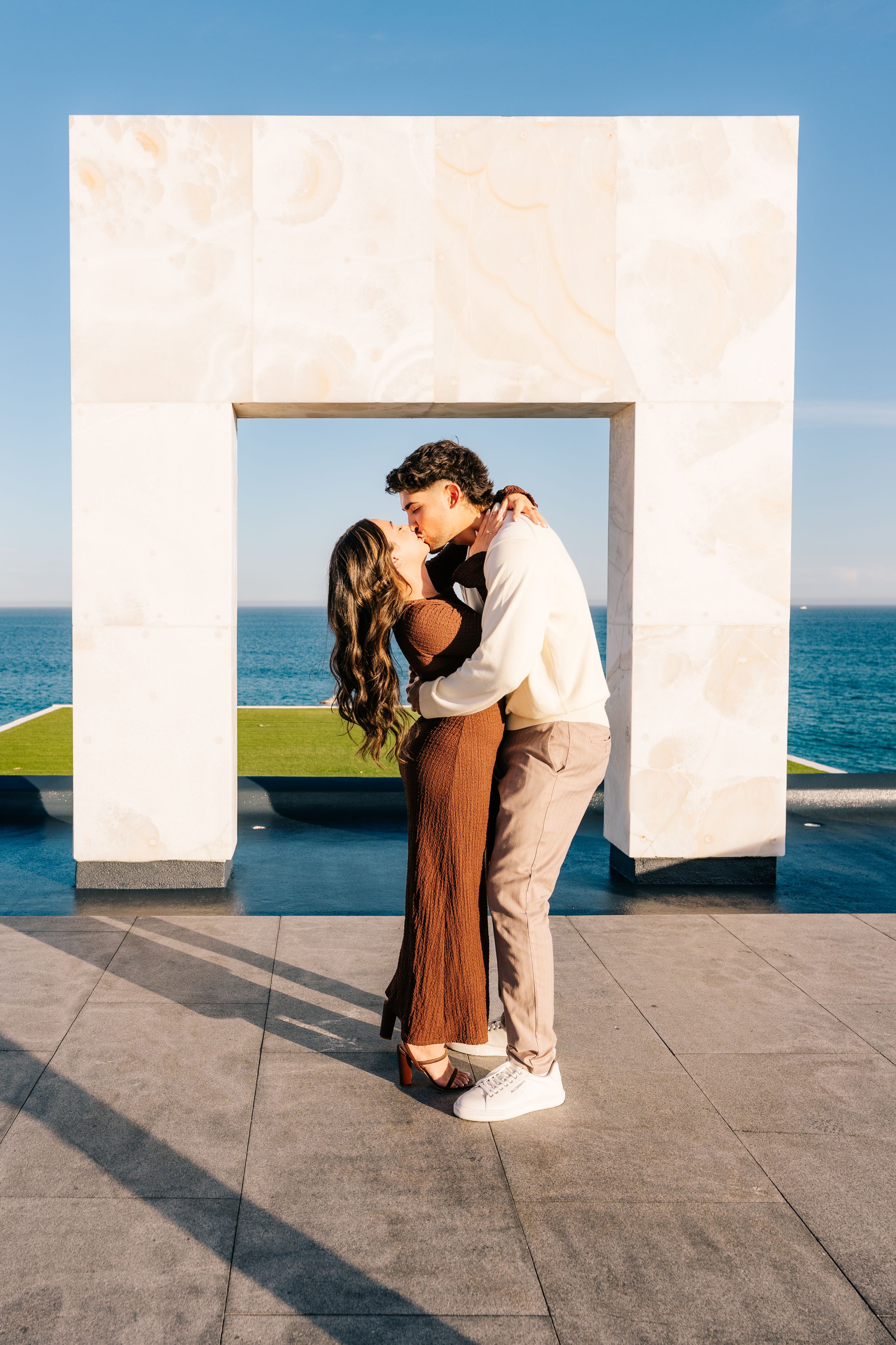 Couple sharing a romantic kiss under a marble arch with ocean views on a sunny day.