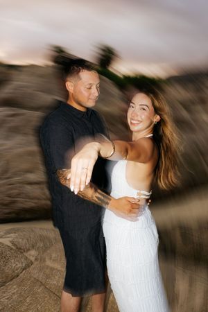 Couple embracing outdoors at dusk, woman in white dress showing engagement ring, man in black outfit with tattoos, motion blur effect.