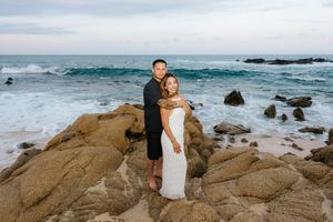 Couple in formal attire posing on rocky beach with ocean waves at sunset.