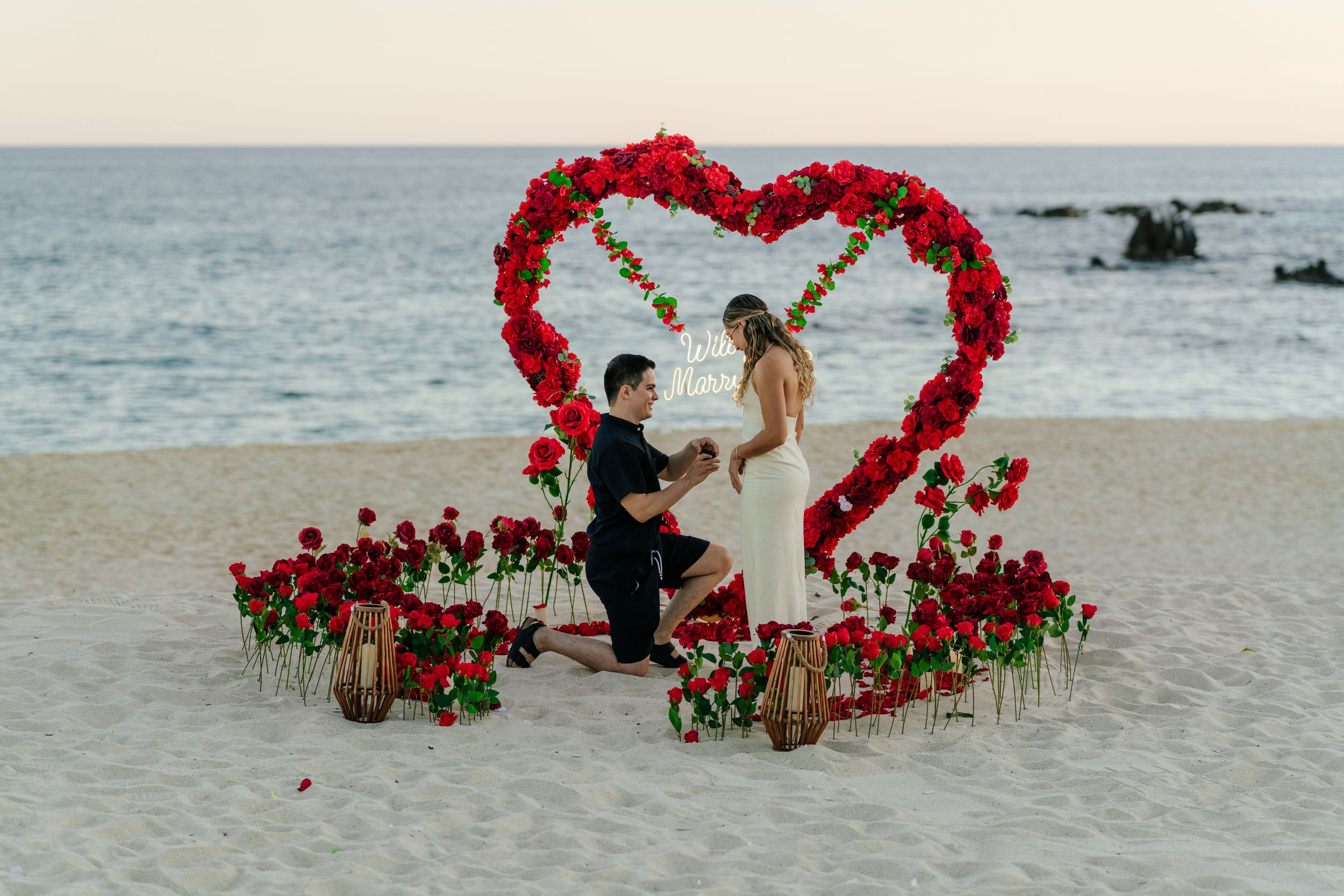 Man proposes to woman on beach under red rose heart arch with rose arrangements on sand at sunset.