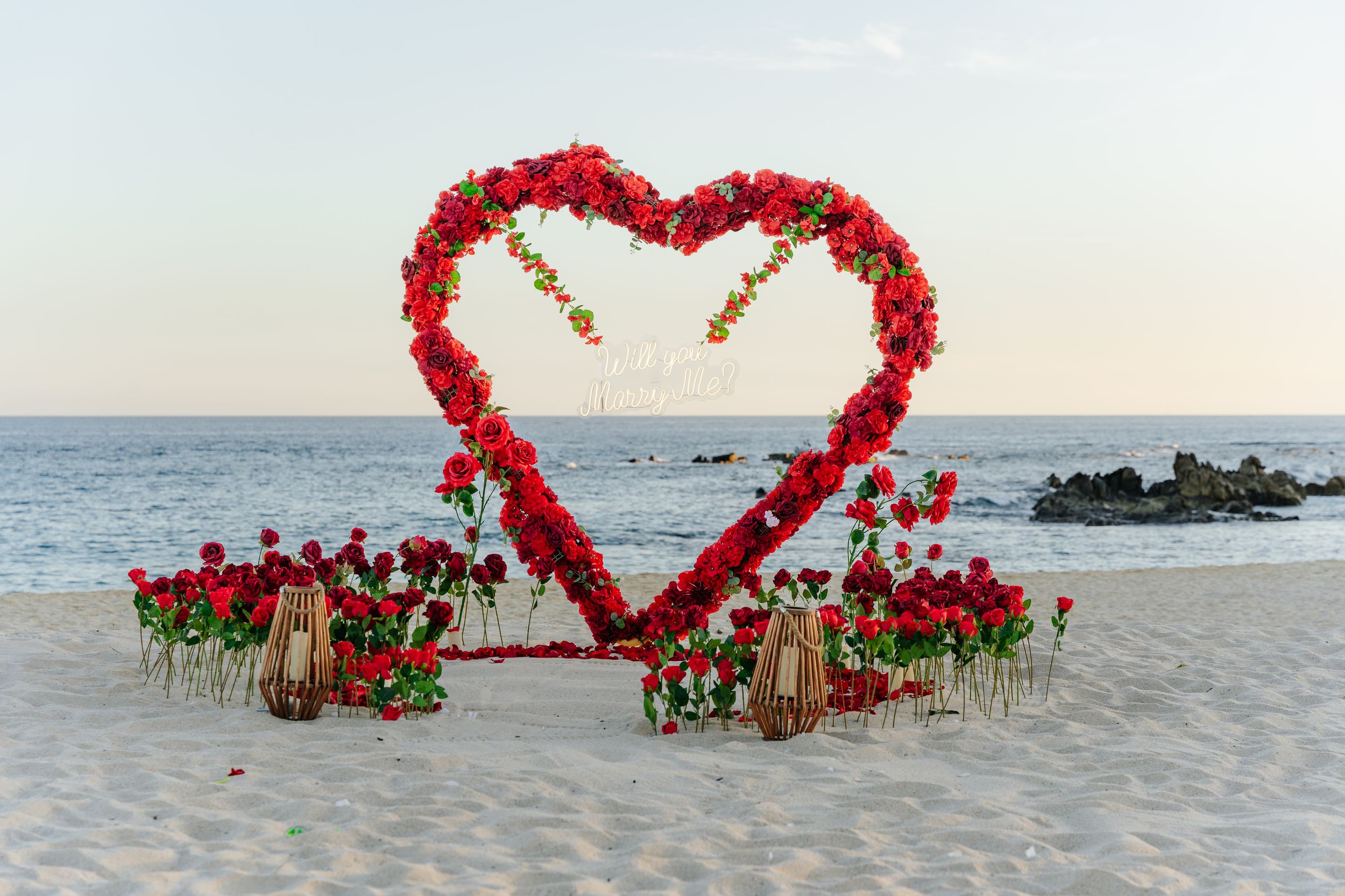 Heart-shaped red floral arch on a sandy beach at sunset, surrounded by rose arrangements and lanterns for a romantic ceremony.