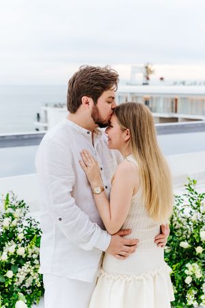 Romantic couple in white outfits sharing a kiss on a seaside terrace surrounded by white floral arrangements.