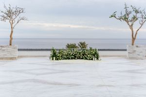 Elegant outdoor wedding ceremony setup with floral arrangement, ocean view, marble floor, and two trees under cloudy sky.