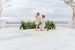 Man proposes on one knee to woman on elegant outdoor terrace with floral arrangements and ocean view backdrop.