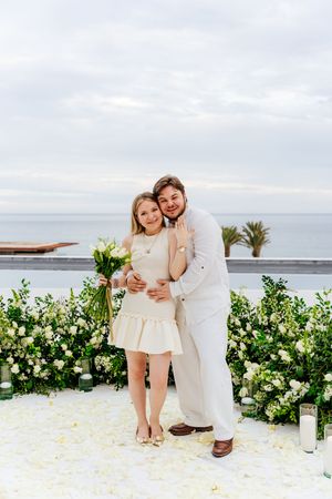 Couple in white attire posing at an outdoor wedding ceremony with white floral arrangements and ocean backdrop.